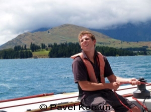 Молодой яхтсмен сидит на шкоте и смотрит за парусом на фоне озера и гор