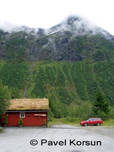 Красный дом с земляной крышей и красный автомобиль на фоне гор поросших деревьями в тумане 
